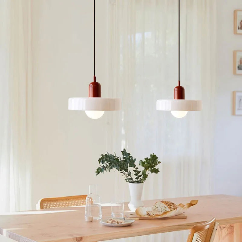 Two modern pendant lights with white ribbed shades and dark red cylindrical accents hang over a light wood dining table in a bright, minimalist room with sheer curtains.