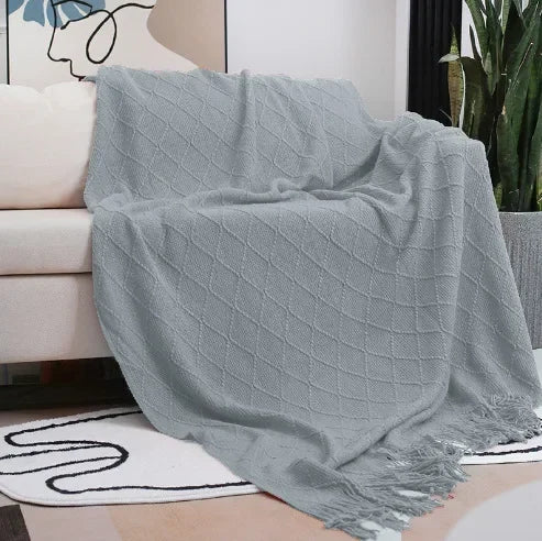 Light grey textured throw with a diamond weave and tassel fringe draped over a sofa arm above a patterned rug