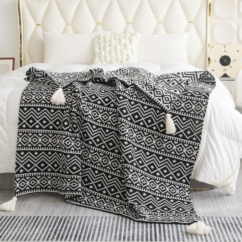 A black-and-off-white patterned throw blanket with cream tassels draped over a bed with white bedding in a bright, modern bedroom.