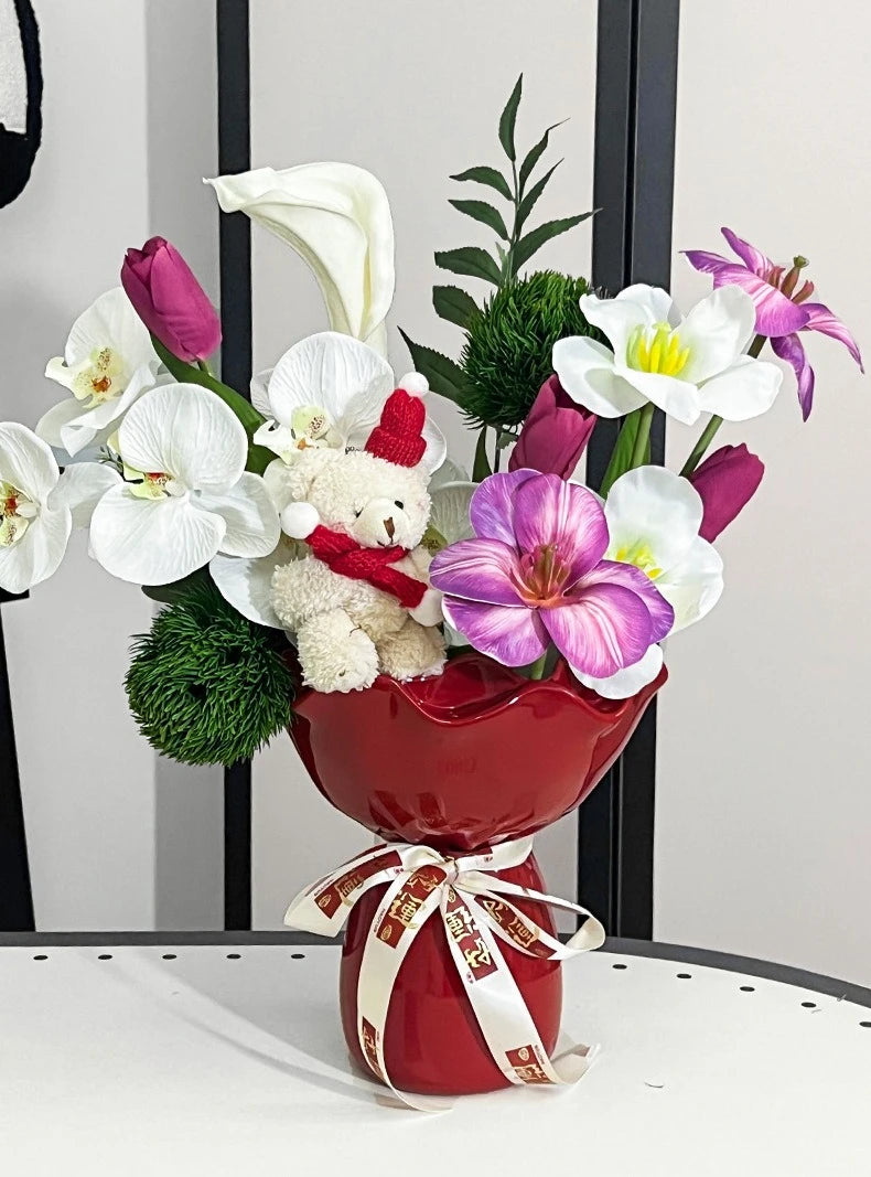 Bouquet of flowers with a teddy bear and a red vase on a white surface.