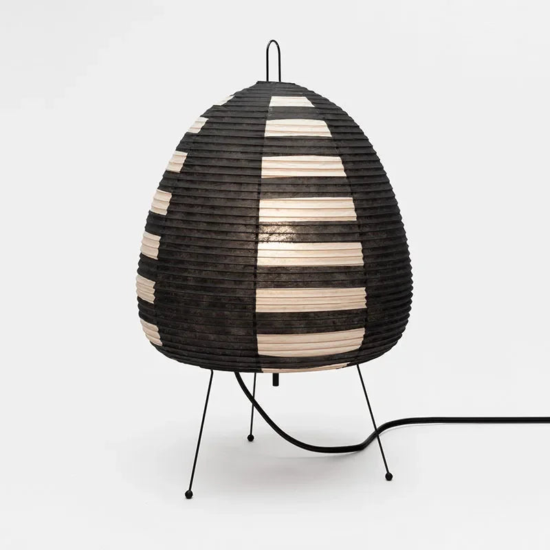 A modern table lamp with a ribbed, egg-shaped shade featuring alternating black and natural beige horizontal stripes, supported by three thin black metal legs on a white background.