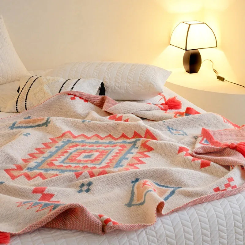 A woven throw blanket with a Southwestern geometric pattern in red, blue, and beige, draped over a white bed in a warm, sunlit bedroom setting.