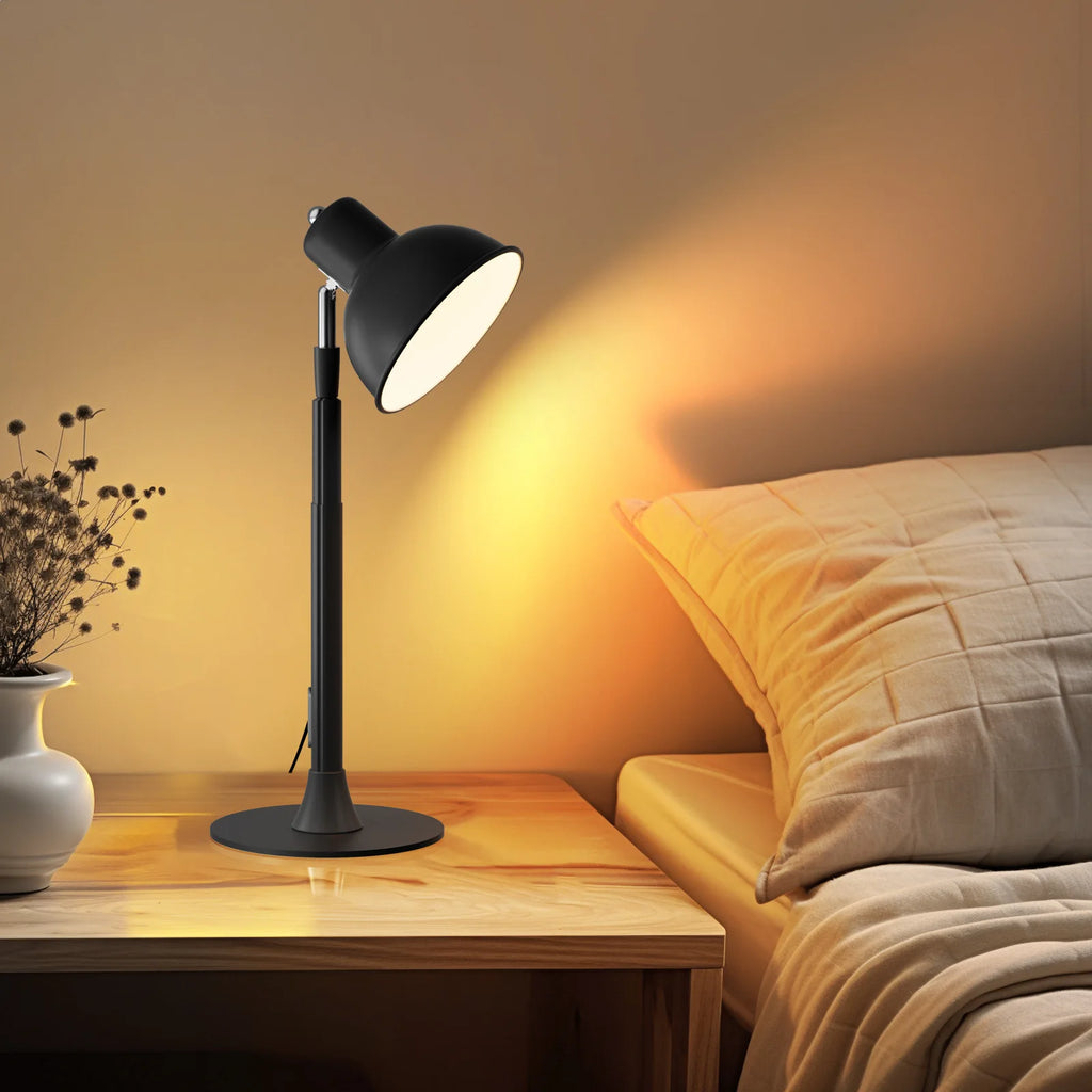 Black table lamp on a wooden nightstand next to a bed with soft lighting.