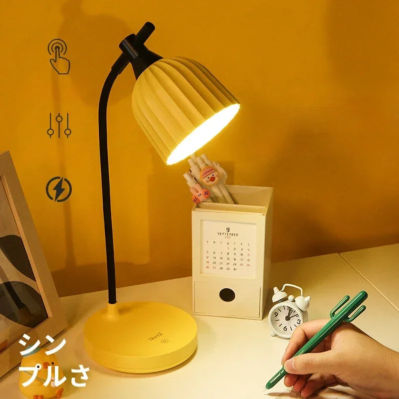 Yellow desk lamp on a table with a hand holding a green pen, against a yellow wall.