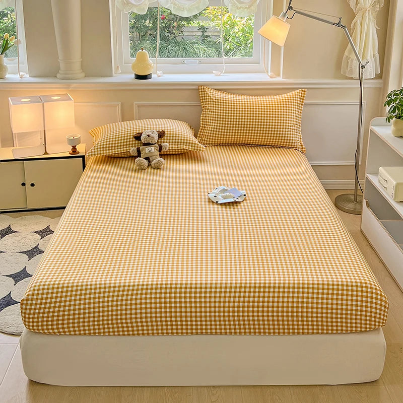 A high-angle shot of a bed featuring a yellow and white mini-plaid gingham fitted sheet and matching pillowcase. A small plush dog toy sits on the pillow against a cream-colored headboard.