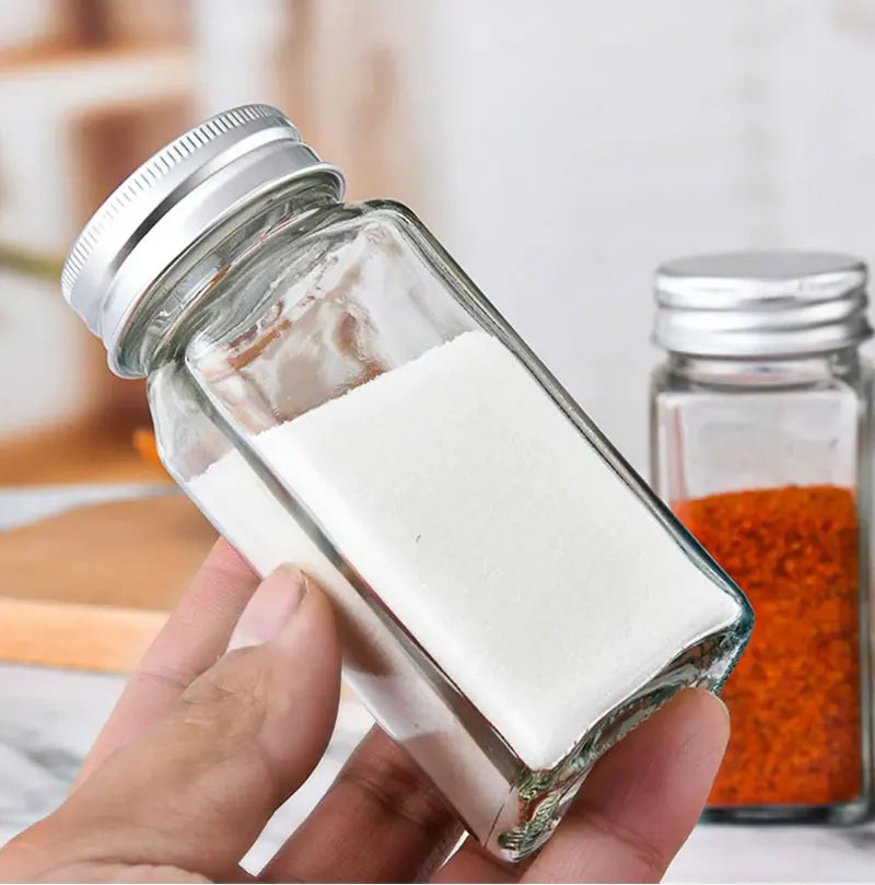 Hand holding a glass jar of white powder with another jar of red powder in the background.
