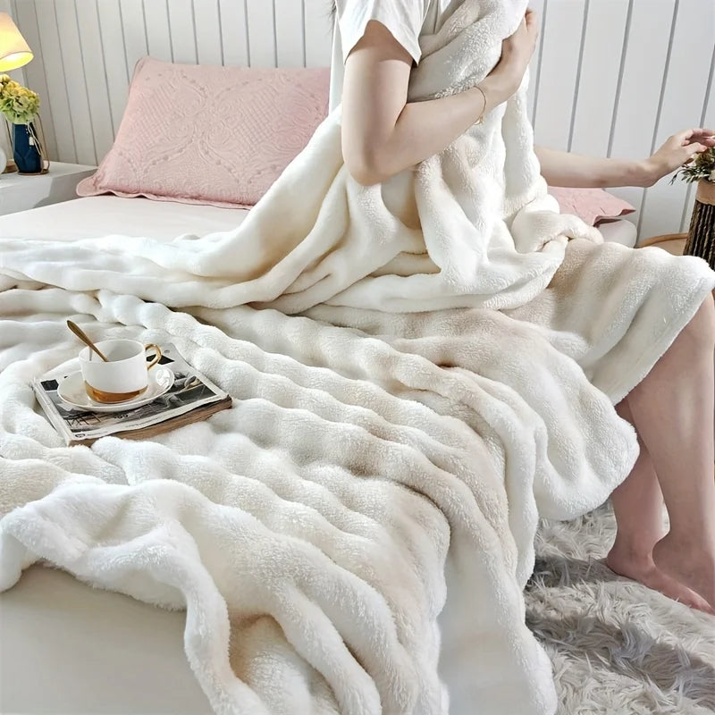 A person sitting on a bed with an ivory white fluffy blanket, holding a cup and a book.