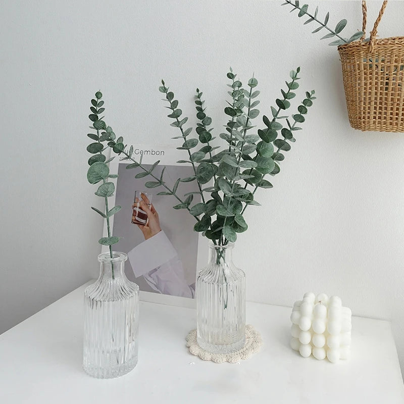 Two glass vases with greenery on a white surface with a neutral background