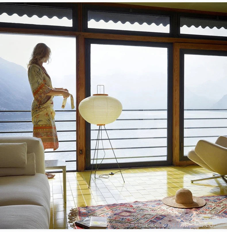 A woman in a room with large windows, standing by a Japanese floor lamp and looking out.