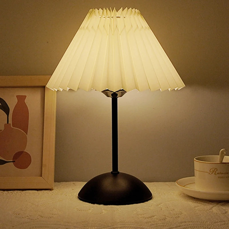 Table lamp with pleated shade on a surface next to a framed picture and cup.