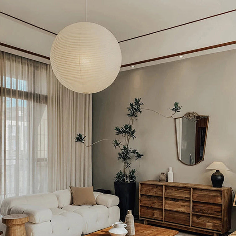 A large spherical white paper lantern pendant light hangs in a modern living room with a white sofa, wooden sideboard, and neutral beige walls.