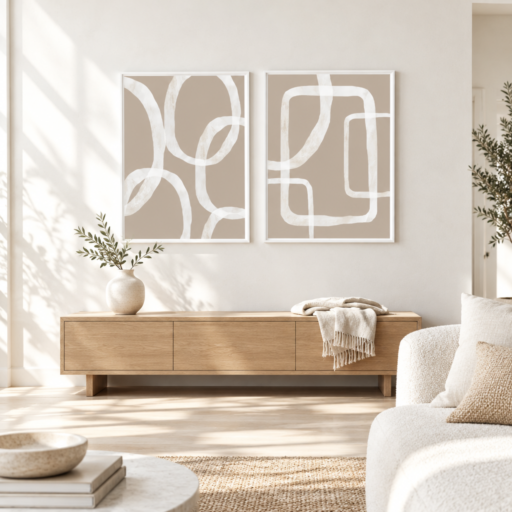 Two framed abstract wall art prints with white curved line patterns on a warm beige background, displayed above a light wood console in a bright, modern living room.