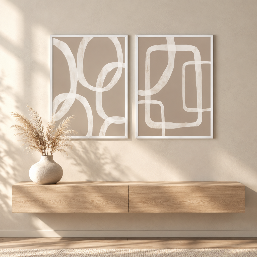 Two vertical framed abstract art prints with white curved line patterns on a warm beige background, displayed side by side on a beige wall above a light wood console table with a ceramic vase of dried grass.