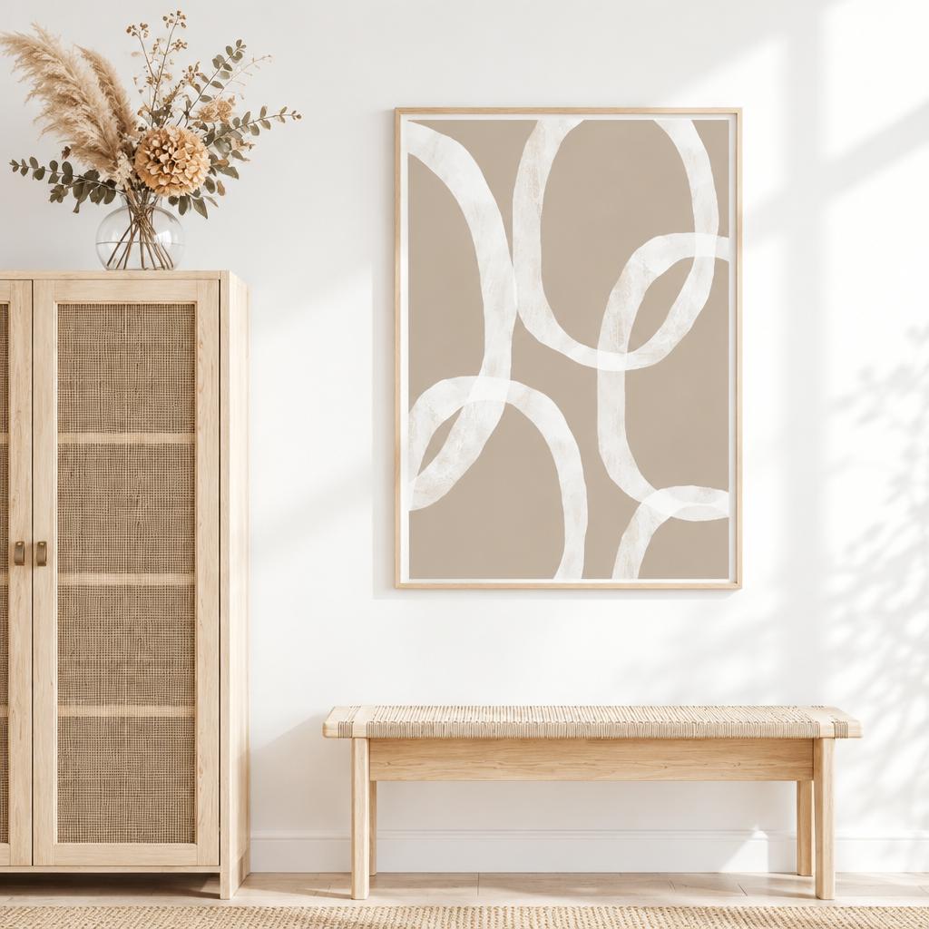 A framed abstract art print with white brushstroke loops on a warm taupe background, displayed on a white wall above a woven bench in a bright, modern room.