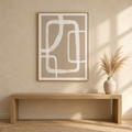 Framed abstract wall art with white curved line shapes on a warm beige background, hanging above a light wood bench in a softly lit interior with a vase of dried grass nearby.